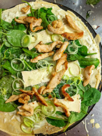 Galettes with Chicken, Spinach, and Brie Cheese Overhead view of Galettes with Chicken, Spinach, and Brie Cheese, with curled leeks and grated cheese scattered over the top on a metal surface.