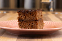 Gingerbread Bars Square Gingerbread Bars with two dark layers and a glossy chocolate top, on a pink ridged plate.