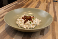 Halušky with Bryndza Cheese Substitution Bowl of Halušky with Bryndza Cheese Substitution topped with crispy bacon strips and herbs, on a wooden counter.