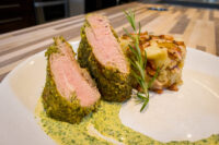 Herb-crusted Pork Tenderloin Sliced Herb-crusted Pork Tenderloin with green herb sauce beside a potato bacon bake and rosemary sprig.