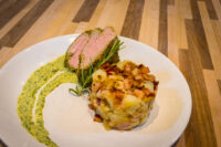 Herb-crusted Pork Tenderloin Sliced Herb-crusted Pork Tenderloin with rosemary, potato hash, and a green herb sauce swirl on a white plate.