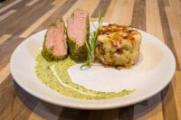 Herb-crusted Pork Tenderloin Sliced Herb-crusted Pork Tenderloin with pink center, rosemary sprig, herb sauce, and potatoes on a white plate
