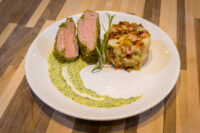 Herb-crusted Pork Tenderloin Sliced Herb-crusted Pork Tenderloin with pink centre, rosemary sprig, potato-bacon hash, and green herb sauce.