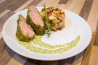 Herb-crusted Pork Tenderloin Sliced Herb-crusted Pork Tenderloin with pink center beside mashed potato and bacon on a swirl of green sauce