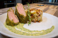 Herb-crusted Pork Tenderloin Sliced Herb-crusted Pork Tenderloin with pink centers beside a potato stack and green sauce on a white plate.