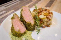 Herb-crusted Pork Tenderloin Sliced Herb-crusted Pork Tenderloin with pink center beside roasted potatoes and rosemary on a white plate.