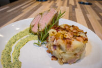 Herb-crusted Pork Tenderloin Sliced Herb-crusted Pork Tenderloin with pink center and herb crust, beside a bacon-topped potato cake.