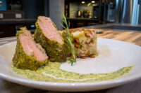 Herb-crusted Pork Tenderloin Sliced Herb-crusted Pork Tenderloin with pink center, beside a golden potato bake and green herb sauce.