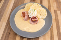 Grey plate with Horseradish Sauce Křenová Omáčka pooled beside rolled ham, cream dollop, and bread rounds.