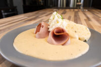 Horseradish Sauce Křenová Omáčka pooled on a grey plate with rolled ham, a cream dollop, and cornbread.