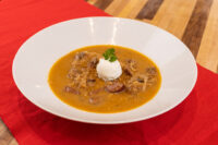 Bowl of Kapustnica Soup with orange broth, meat chunks, shredded cabbage, sour cream, and parsley.