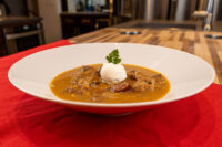 White bowl of orange-brown Kapustnica Soup with meat chunks, cream dollop, and parsley on red cloth.