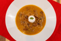 Bowl of Kapustnica Soup with sausage, meat, and cabbage strands in orange broth, topped with cream and herb.