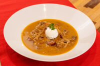 White bowl of orange-brown Kapustnica Soup with sausage chunks, cream dollop, and parsley garnish on red cloth.