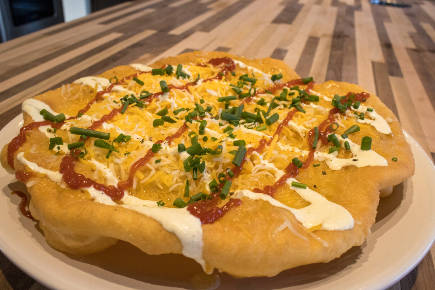 Langos Langos flatbread on a white plate topped with sour cream, red sauce, shredded cheese, and chives.