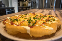 Langos Langos: puffed fried dough topped with melted cheese, red sauce, white sauce drizzle, and chopped chives on a plate.