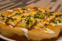 Langos Fried Langos flatbread topped with sour cream, red sauce, shredded cheese, and chives on a white plate.