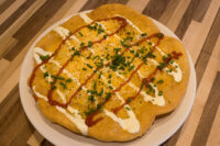 Langos Golden fried Langos flatbread topped with cheese, chives, and red and white sauce drizzles on a plate.