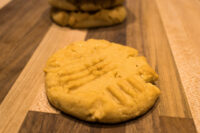 Peanut Butter Cookies A Peanut Butter Cookie with crosshatch fork marks on a wooden board, more cookies stacked behind it.