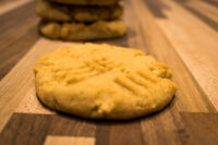 Peanut Butter Cookies A golden Peanut Butter Cookie with crosshatch fork marks, more cookies stacked behind it on a wooden table.