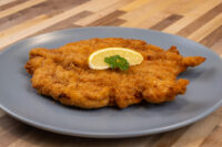 Golden fried Chicken Schnitzel on a grey plate, topped with a lemon wedge and parsley sprig.