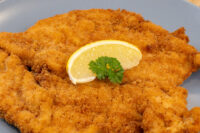 Golden breaded Chicken Schnitzel on a gray plate with a lemon wedge and parsley garnish.