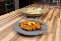Golden fried chicken schnitzel with lemon slice on a grey plate, next to a bowl of creamy salad.