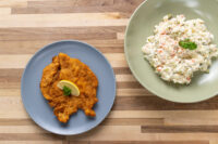 Golden Chicken Schnitzel with a lemon wedge on a grey plate beside a bowl of potato salad.
