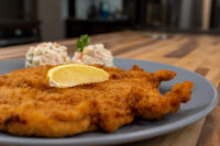 Golden fried Chicken Schnitzel on a grey plate with a lemon slice and creamy salad garnish.
