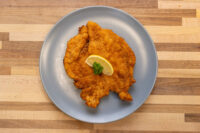 Golden breaded Chicken Schnitzel on a grey plate, garnished with a lemon slice and fresh herb sprig.