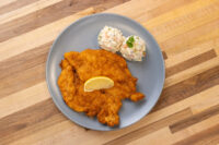 Golden breaded Chicken Schnitzel on a grey plate with a lemon wedge and creamy potato salad.