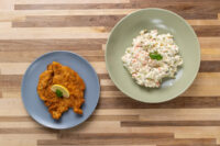 Golden Chicken Schnitzel with lemon wedge on a grey plate beside creamy potato salad on wood table.