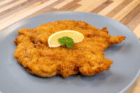 Golden fried Chicken Schnitzel on a grey plate, topped with a lemon wedge and parsley garnish.