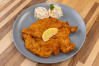 Golden breaded Chicken Schnitzel on a grey plate with a lemon wedge and creamy potato salad.
