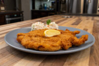 Golden fried Chicken Schnitzel on a gray plate with a lemon wedge, herb sprig, and coleslaw