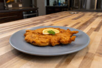Golden fried Chicken Schnitzel on a gray plate with a lemon slice and herb garnish on a wooden counter.