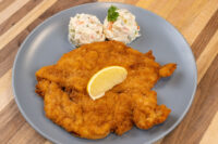 Golden breaded Chicken Schnitzel with a lemon wedge, served beside creamy potato salad on a gray plate.