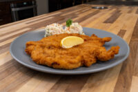 Golden breaded Chicken Schnitzel on a grey plate with a lemon wedge and potato salad garnished with parsley.