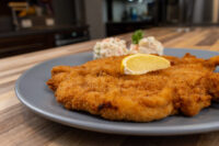 Golden fried Chicken Schnitzel topped with a lemon wedge, served with a creamy salad on a grey plate.