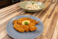 Golden breaded Chicken Schnitzel on a grey plate with lemon wedge and herb, beside a bowl of creamy salad.