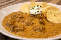 Bowl of Szegedin Goulash with sour cream and chives, served with bread rolls on a wooden table.