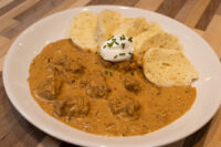 Bowl of Szegedin Goulash with sour cream, chives, and bread slices on a white plate.