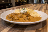 Plate of Szegedin Goulash in creamy paprika sauce, topped with sour cream and chives, with bread wedges.