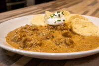 Plate of Szegedin Goulash with creamy paprika sauce, sour cream, chives, and bread slices on a wooden table.