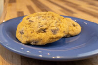 Chocolate Chip Cookies Two golden Chocolate Chip Cookies with visible chips, resting on a blue plate atop a wooden table.