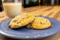 Chocolate Chip Cookies Two Chocolate Chip Cookies on a blue plate with a glass of milk behind them on a wooden table.