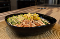 Poke Bowl with Salmon Black bowl of Poke Bowl with Salmon topped with avocado, mango, cucumber, and spicy mayo drizzle.
