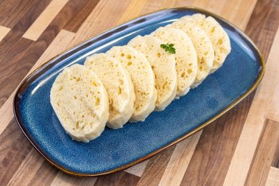 Czech Bread Dumpling Houskovy Knedlik Recipe Sliced houskový knedlík rounds with airy texture on a blue oval plate, garnished with herb.