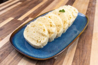 Czech Bread Dumpling Houskovy Knedlik Recipe Sliced houskový knedlík rounds with airy crumb on a blue oval plate, garnished with parsley.