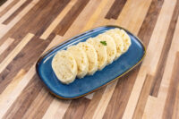Czech Bread Dumpling Houskovy Knedlik Recipe Sliced rounds of houskový knedlík arranged on a blue oval plate with a herb garnish.